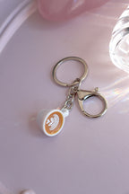 Miniature white latte cup keychain with latte art, a silver-tone key ring and lobster clasp on a pink background.